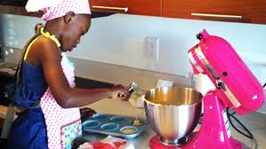 Black woman in a pink apron mixes baking batter at a kitchen aid as Marie Deveaux, business coach describes how small business owners who are still working it as a side hustle can still make baby steps towards big goals.