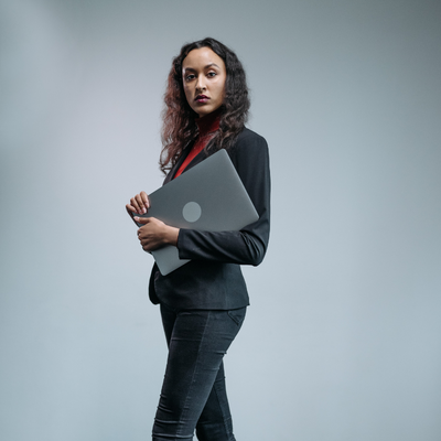 A woman in business attire is smiling and waiting while leaning against a pole.