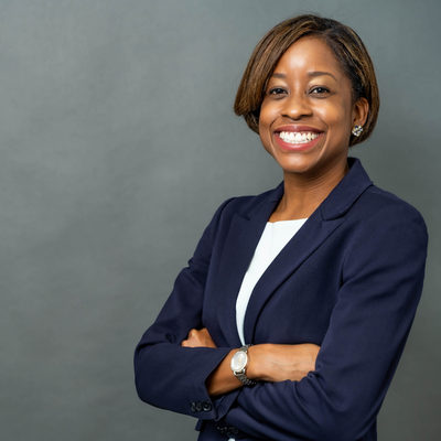 A woman in business attire is smiling and waiting while leaning against a pole.